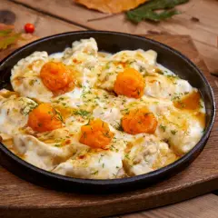 Pierożki z dynią w sosie, posypane koperkiem, podane w żeliwnej patelni na drewnianym blacie. Obok kawałek dyni.