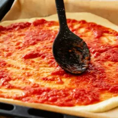 Rozsmarowywanie sosu do pizzy na cieście za pomocą czarnej łyżki.