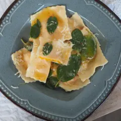 Pyszne ravioli z dynią, polane masłem z szałwią i posypane parmezanem, na niebieskim talerzu.