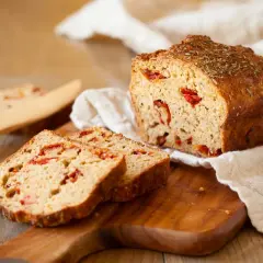 Chleb z suszonymi pomidorami, pokrojony na kromki, leży na desce. Wierzch bochenka posypany ziołami.