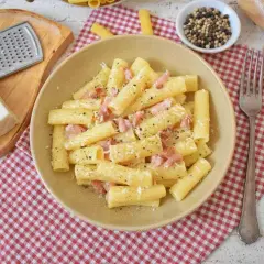 Pyszne rigatoni carbonara z boczkiem i parmezanem, posypane świeżo mielonym pieprzem. Obok tarka i kawałek sera.
