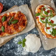 Pyszna pinsa z burratą, rukolą i pomidorkami. Obok druga pinsa z salami i bazylią, ciasto i pomidor.