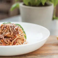 Pyszne spaghetti z bogatym sosem bolońskim, posypane parmezanem, podane na białym talerzu. Obok kawałek sera i listek bazylii.