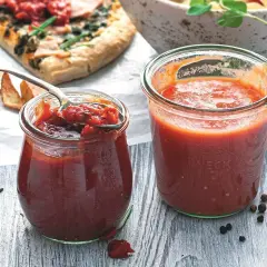 Dwa słoiki sosu pomidorowego, jeden z łyżką, obok pizzy i przypraw.