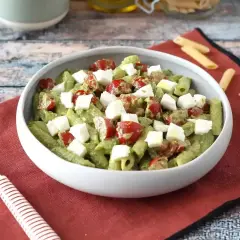 Pyszna pasta pesto z pomidorkami koktajlowymi i kostkami mozzarelli w białej misce, na czerwonej serwetce.