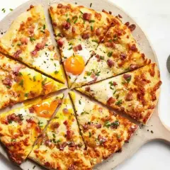 Kawałki pizzy carbonara z jajkiem sadzonym, boczkiem i szczypiorkiem na desce.