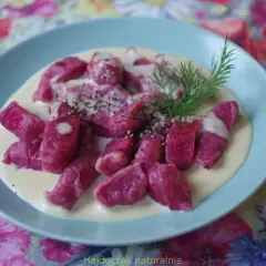Różowe gnocchi buraczane w kremowym sosie, posypane pieprzem i udekorowane gałązką koperku, na talerzu z kwiecistym obrusem.