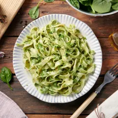 Pyszne zielone tagliatelle z pesto, podane na talerzu z niebieskimi paskami. Obok garnek z pieprzem, czosnek i oliwa.