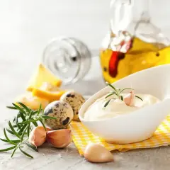 Kremowy sos aioli w białej miseczce, ozdobiony gałązką rozmarynu i ząbkiem czosnku. Obok składniki: jajka przepiórcze, cytryna, czosnek i oliwa.