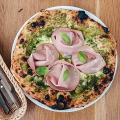 Pizza z mortadelą i pistacjami, zielonym pesto i listkami bazylii. Obok koszyk z widelcem i nożem.