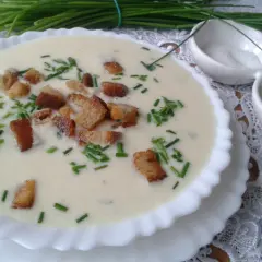 Kremowa zupa porowa z grzankami i szczypiorkiem, podana w białej miseczce. Obok małe miseczki z solą i pieprzem.