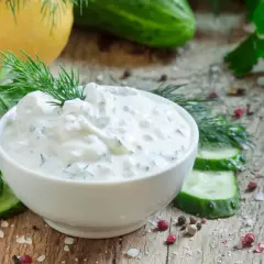 Pyszny dip jogurtowy z ziołami i ogórkiem, udekorowany koperkiem, podany z plastrami ogórka i przyprawami na drewnianym blacie.