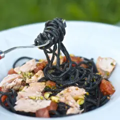 Czarny makaron z łososiem, pomidorkami i pistacjami, nabierany na widelec z czerwoną rączką.