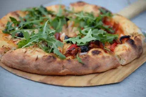 Pyszne ciasto na pizzę z rukolą, oliwkami i papryką, podane na drewnianej desce. Idealne na letni wieczór.