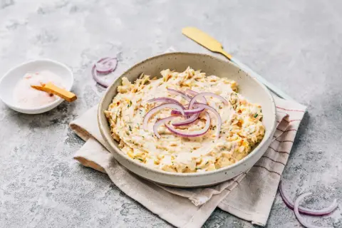 Kremowa pasta z tuńczyka, udekorowana krążkami czerwonej cebuli, podana w misce na lnianej serwetce. Obok miseczka z różową solą.