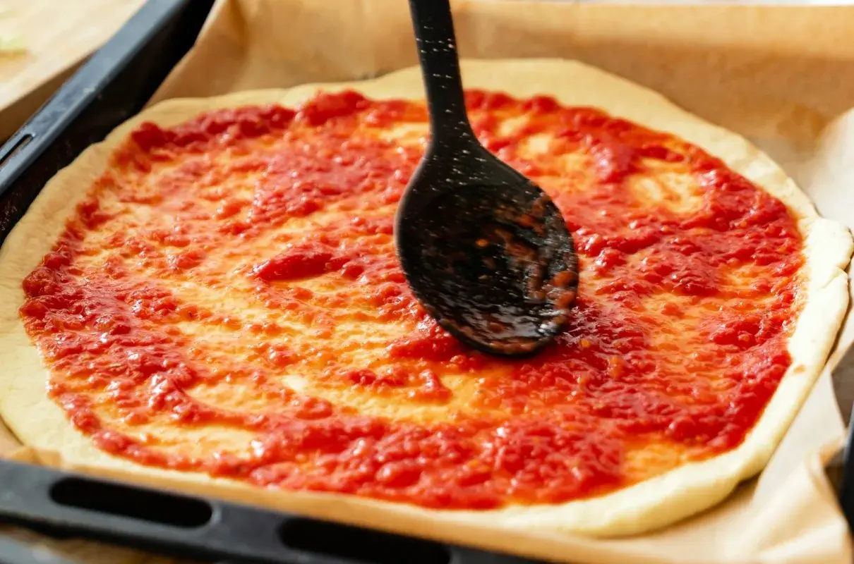 Rozsmarowywanie sosu do pizzy na cieście za pomocą czarnej łyżki.