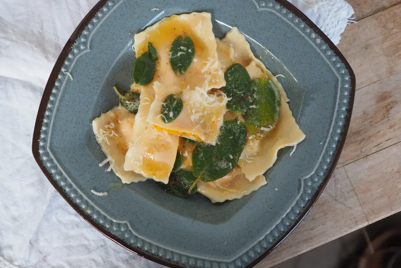 Pyszne ravioli z dynią, polane masłem z szałwią i posypane parmezanem, na niebieskim talerzu.