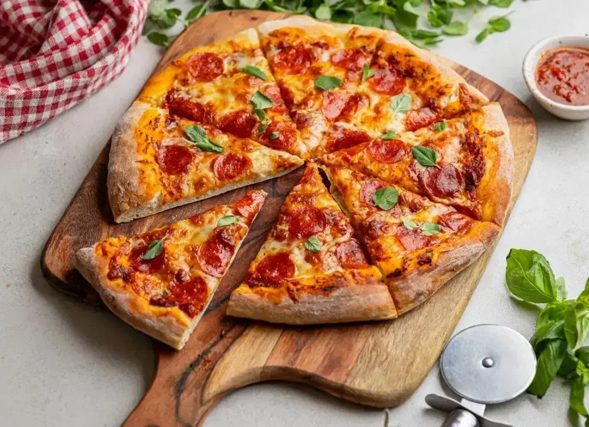 Pyszna pizza z pepperoni i świeżą bazylią, pokrojona na kawałki, leży na drewnianej desce. Obok sos pomidorowy i nóż do pizzy.