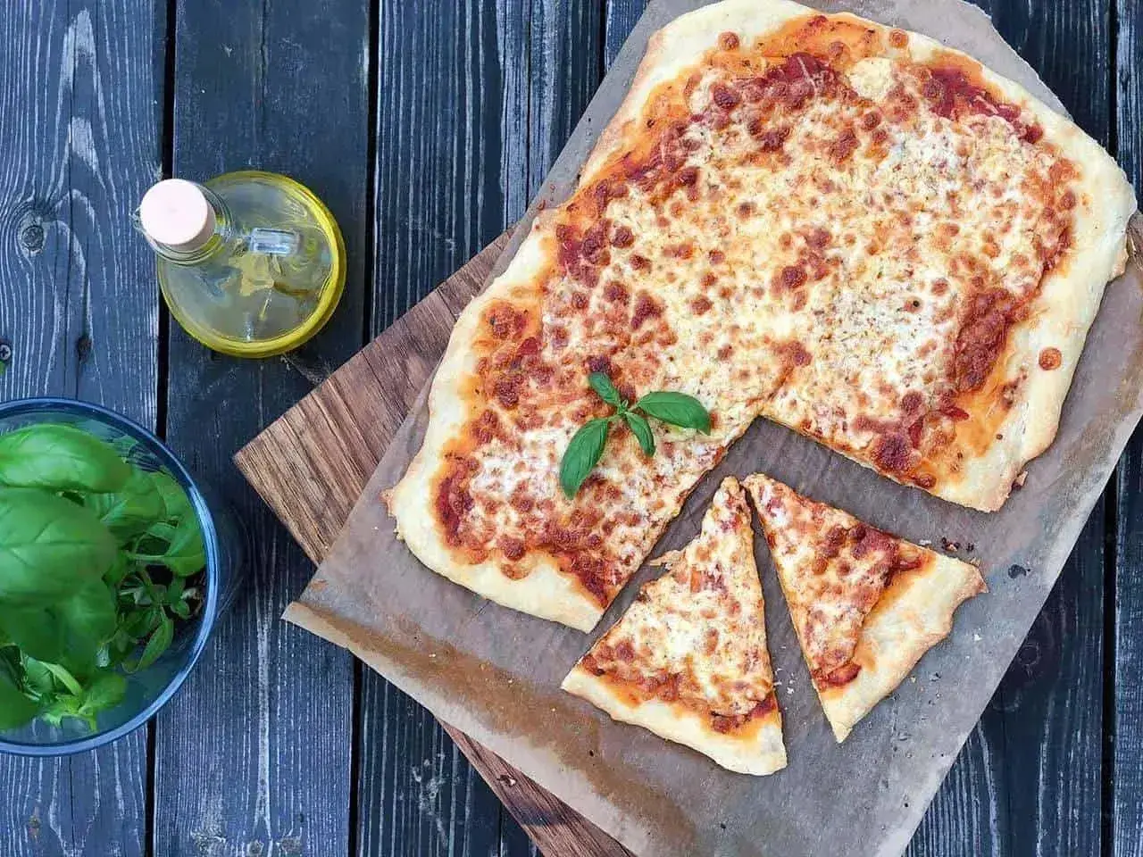 Pyszna pizza margherita z roztopionym serem i świeżą bazylią, obok buteleczka oliwy i miseczka z ziołami.