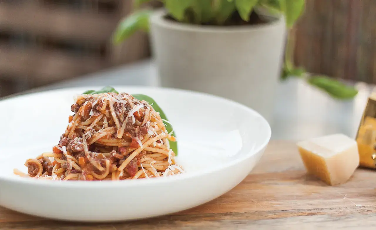 Pyszne spaghetti z bogatym sosem bolońskim, posypane parmezanem, podane na białym talerzu. Obok kawałek sera i listek bazylii.