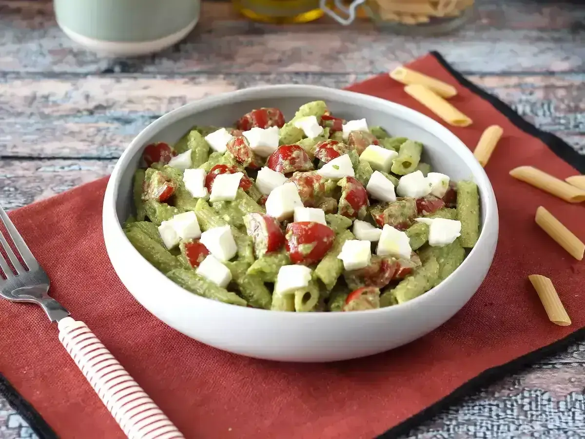 Pyszna pasta pesto z pomidorkami koktajlowymi i kostkami mozzarelli w białej misce, na czerwonej serwetce.