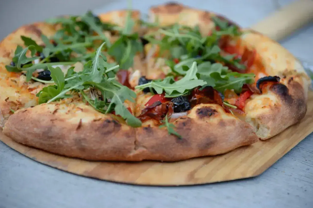 Pyszne ciasto na pizzę z rukolą, oliwkami i papryką, podane na drewnianej desce.