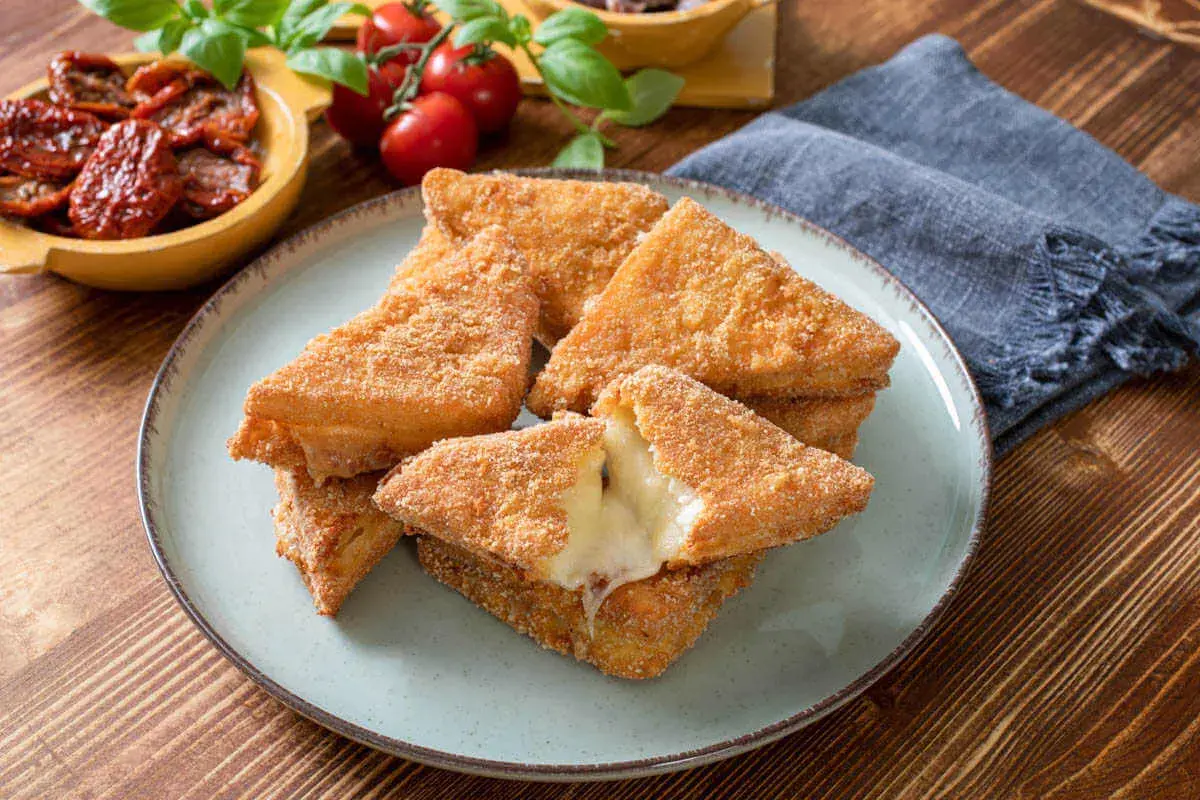Chrupiące mozzarella in carrozza, z rozpływającym się serem w środku, na talerzu. Obok pomidorki i suszone pomidory.