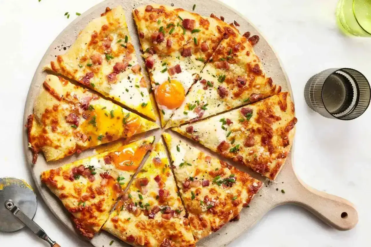 Kawałki pizzy carbonara z jajkiem sadzonym, boczkiem i szczypiorkiem na desce.