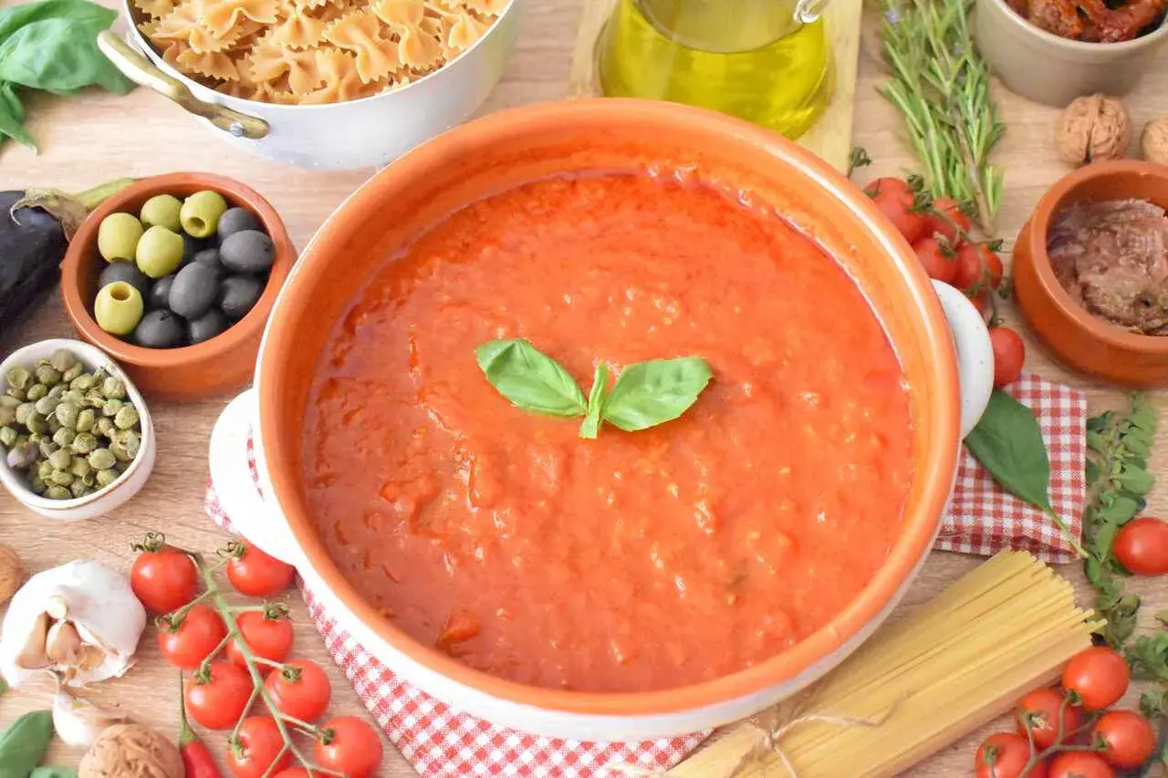 Aksamitny sos al pomodoro w glinianej misie, otoczony składnikami: makaronem, oliwkami, kaparami, pomidorkami, czosnkiem i bazylią.