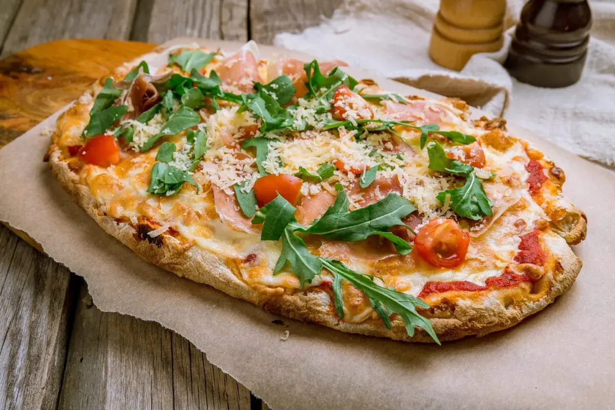 Pyszna pinsa romana z rukolą, pomidorkami i szynką parmeńską, posypana parmezanem, na drewnianym stole.