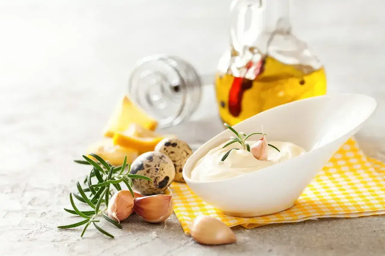 Kremowy sos aioli w białej miseczce, ozdobiony gałązką rozmarynu i ząbkiem czosnku. Obok składniki: jajka przepiórcze, cytryna, czosnek i oliwa.