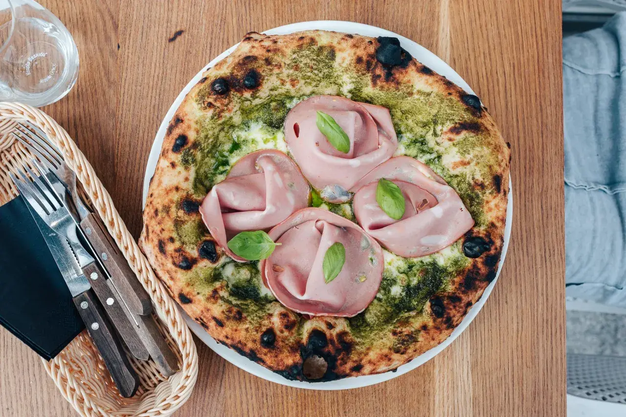 Pizza z mortadelą i pistacjami, zielonym pesto i listkami bazylii. Obok koszyk z widelcem i nożem.