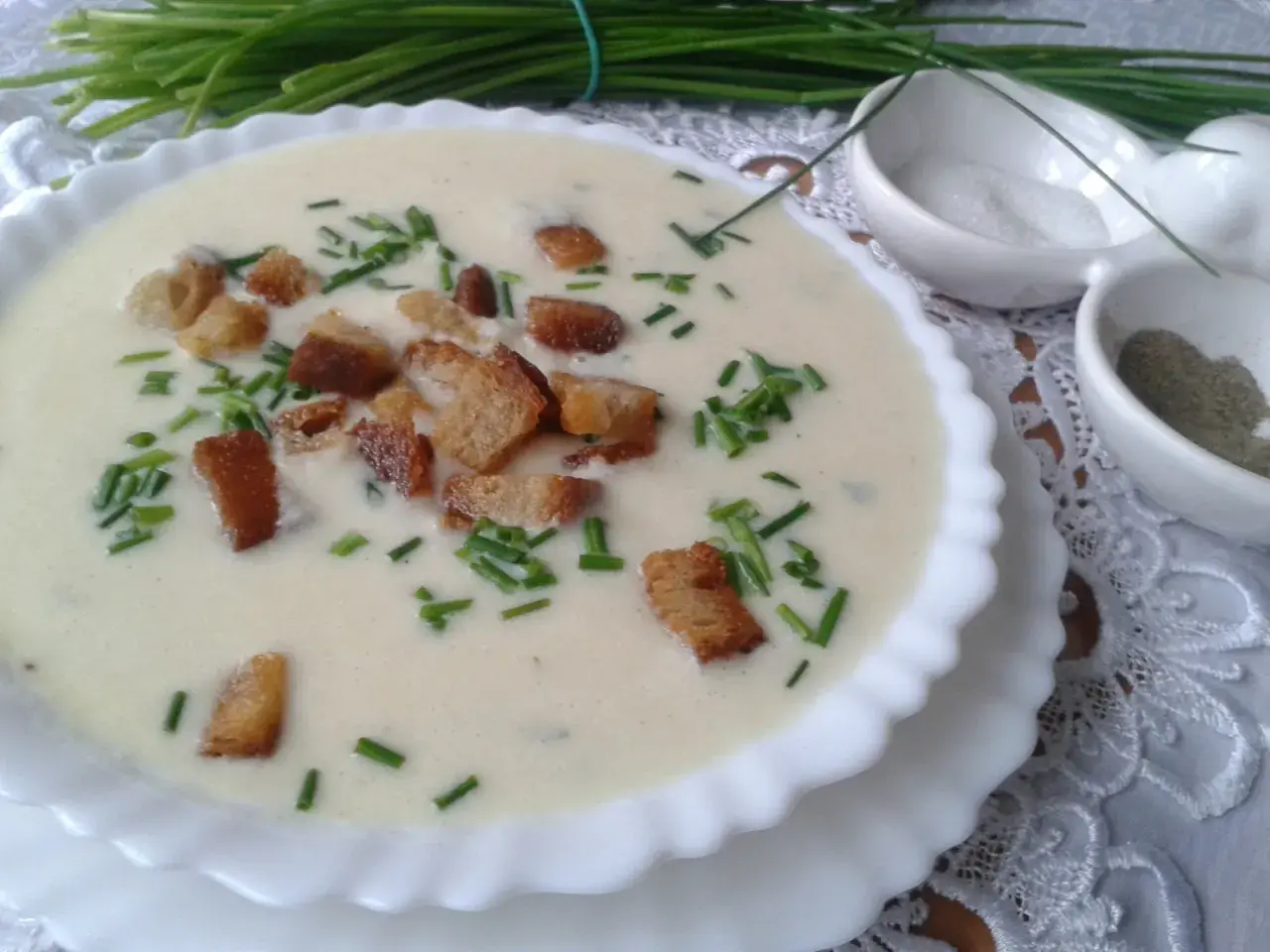 Kremowa zupa porowa z grzankami i szczypiorkiem, podana w białej miseczce. Obok małe miseczki z solą i pieprzem.