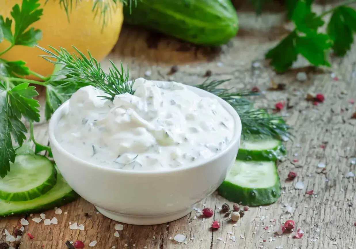 Pyszny dip jogurtowy z ziołami i ogórkiem, udekorowany koperkiem, podany z plastrami ogórka i przyprawami na drewnianym blacie.