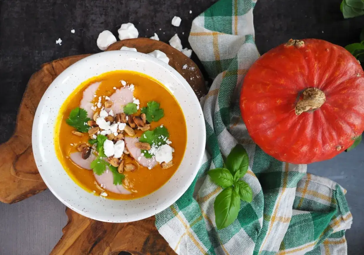 Aksamitna zupa dyniowa z kawałkami mięsa, serem feta, orzeszkami i świeżą kolendrą. Obok leży cała pomarańczowa dynia.