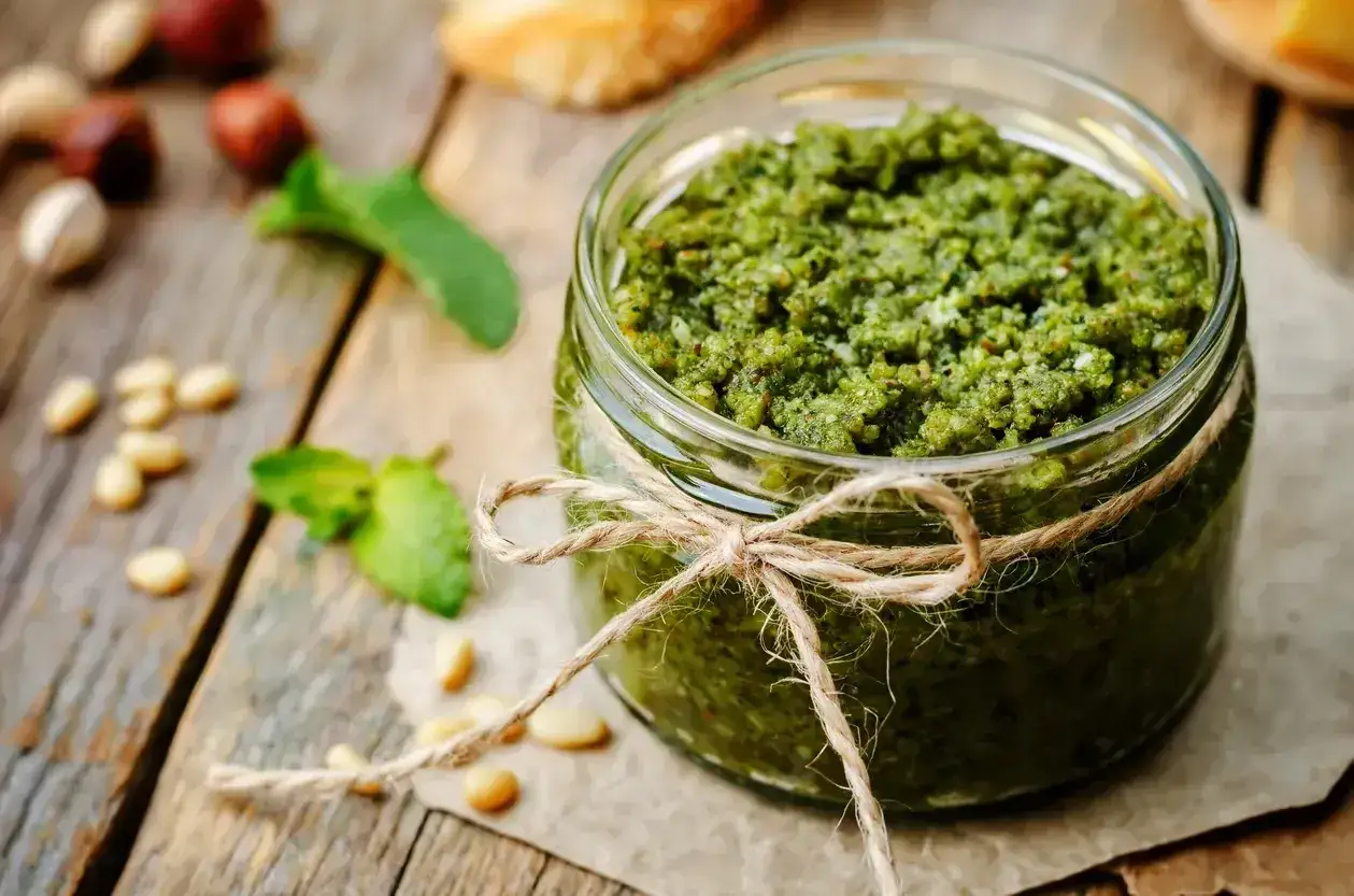 Słoik domowego pesto bazyliowego, ozdobiony sznurkiem, na drewnianym blacie z orzeszkami pinii i listkami bazylii.
