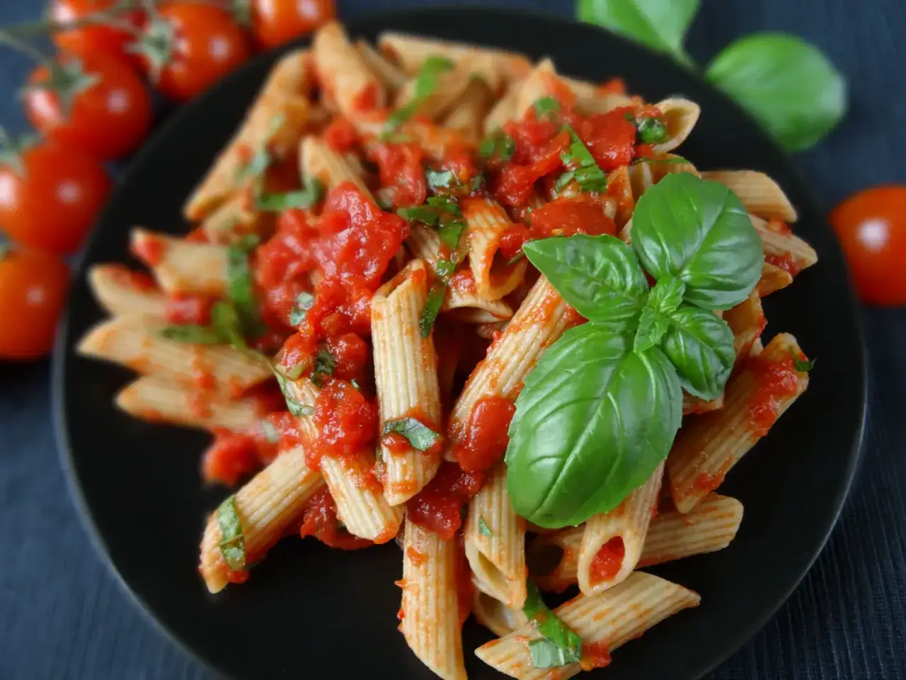 Penne rigate z sosem pomidorowym i świeżą bazylią na czarnym talerzu. Obok kilka pomidorków koktajlowych.