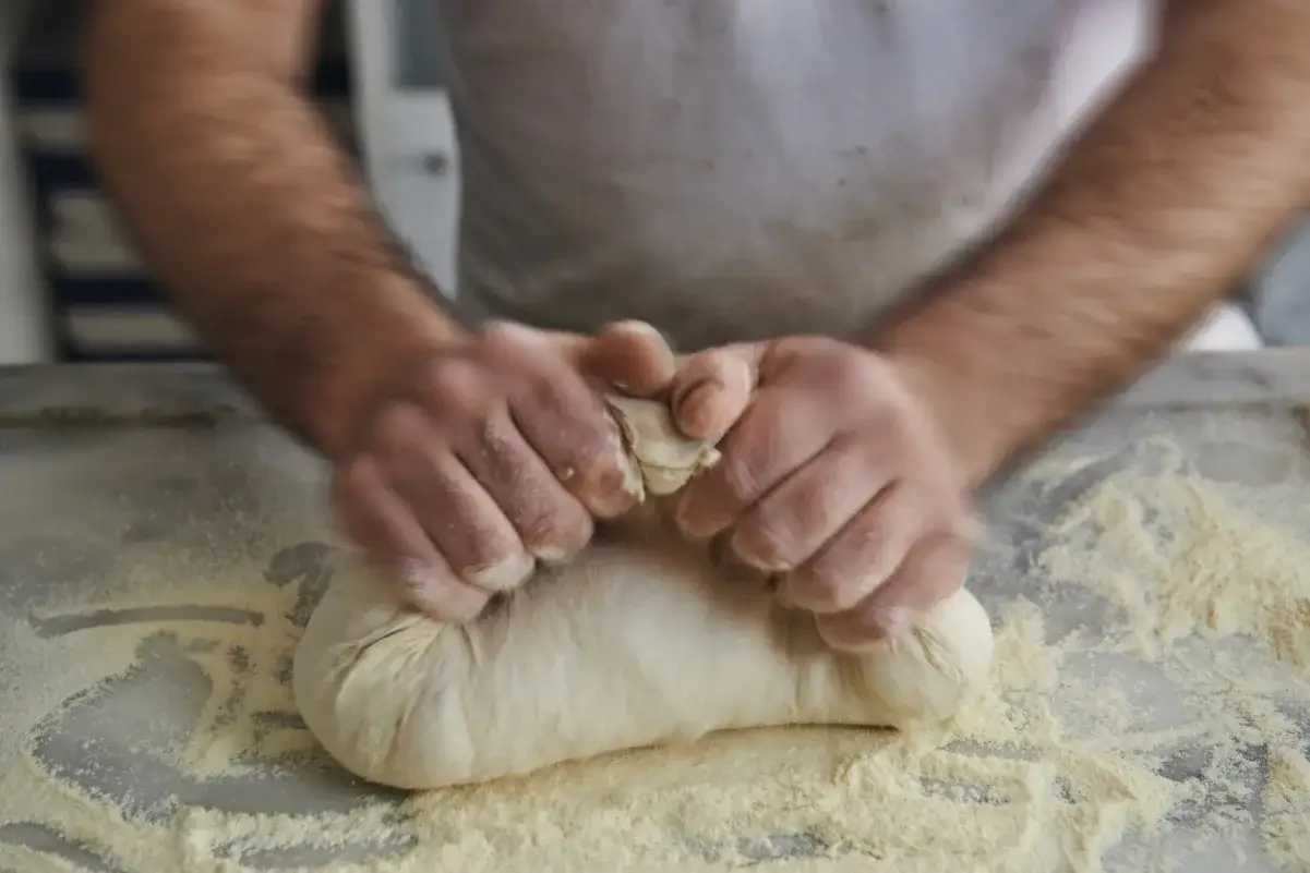 Dłonie ugniatające puszyste ciasto na pizzę, obsypane mąką.