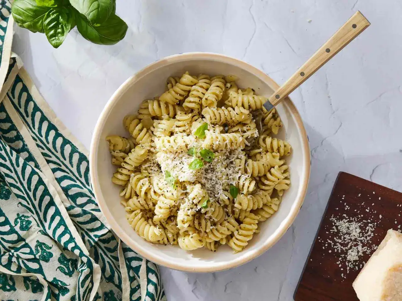Miseczka makaronu fusilli z pesto, posypana startym serem. Obok kawałek parmezanu i świeża bazylia.