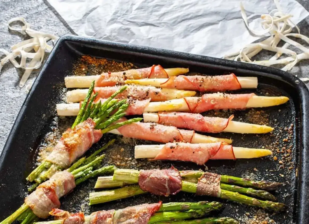 Zielone i białe szparagi owinięte w szynkę parmeńską, ułożone na tacy, posypane przyprawami.