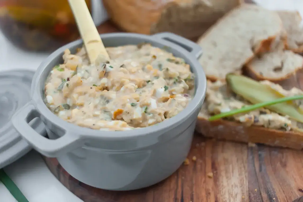 Pyszna pasta z tuńczyka z jajkiem i korniszonami, podana w szarej miseczce, obok kromek chleba.