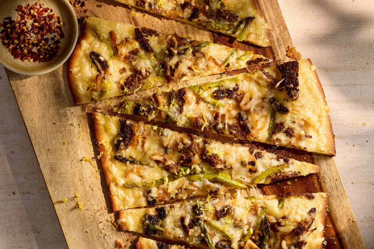 Pyszna pizza ze szparagami i roztopionym serem, pokrojona na kawałki, czeka na desce. Obok miseczka z płatkami chili.