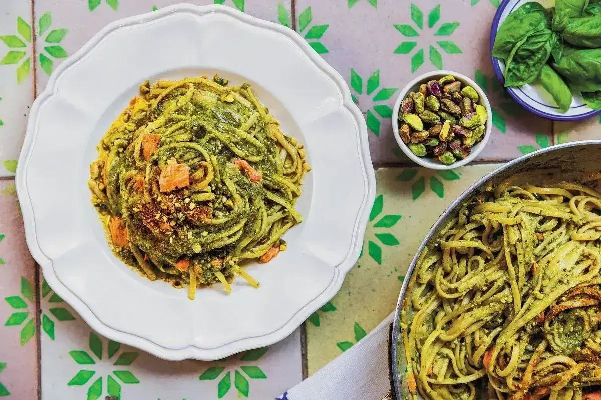 Pyszne spaghetti z zielonym pesto, posypane pistacjami i kawałkami suszonych pomidorów. Obok miseczka z pistacjami i listkami bazylii.