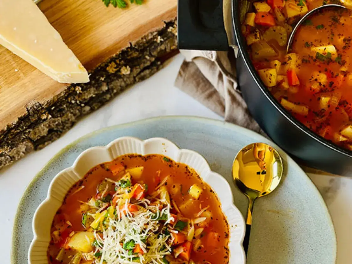 Aksamitna zupa minestrone z warzywami i parmezanem, podana w ozdobnej misce, obok kawałka sera na drewnianej desce.