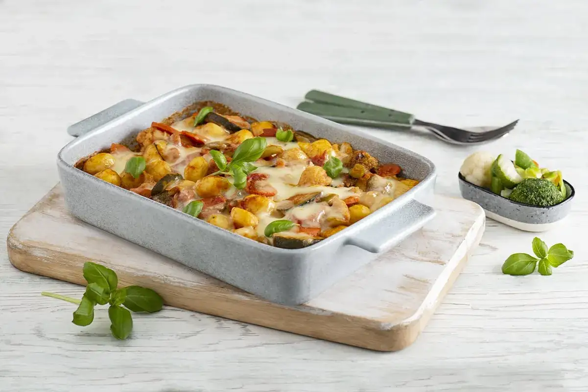 Pyszne zapiekane gnocchi z warzywami i ciągnącym się serem, udekorowane listkami bazylii. Obok miseczka z gotowanymi warzywami.