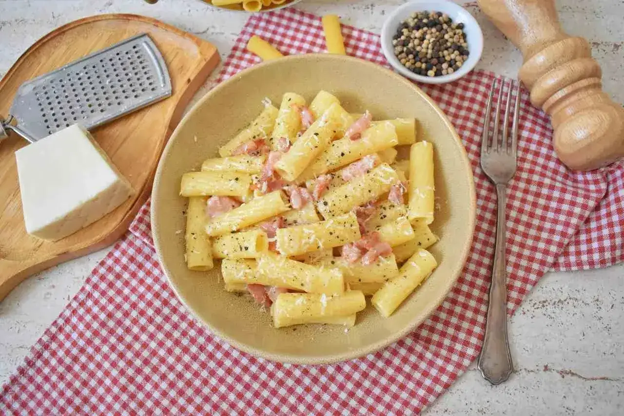 Pyszne rigatoni carbonara z boczkiem i parmezanem, posypane świeżo mielonym pieprzem. Obok kawałek sera i tarka.
