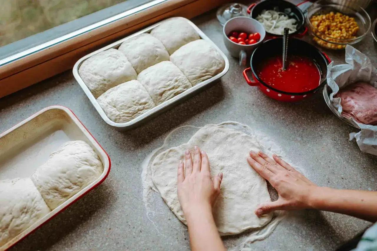 Ręce formują ciasto na pizzę, obok rosnące bułeczki i składniki. Proste ciasto na pizzę zawsze wychodzi!
