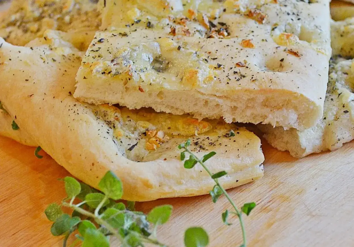 Puszysta focaccia z mozzarellą, posypana ziołami i pieprzem, prezentuje się apetycznie na drewnianej desce.