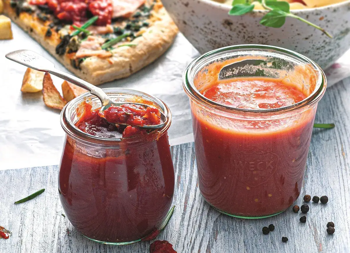 Dwa słoiki domowego sosu pomidorowego do pizzy. Jeden otwarty, z łyżką pełną gęstego sosu, obok pizza i pieczone ziemniaki.