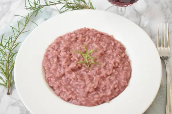 Aksamitne risotto z czerwonym winem, udekorowane gałązką rozmarynu, prezentuje się apetycznie na białym talerzu. Obok widoczny widelec i kieliszek wina.
